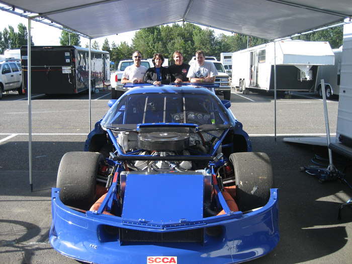 Specialty Engineering blue GT1 Camaro with hood off and racing engine exposed. Andy Pearson, Tracey Pearson and their crew pose behind the race car